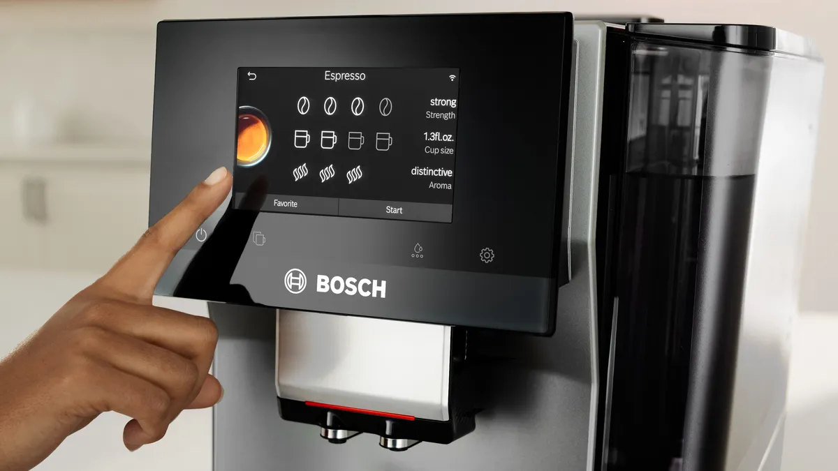 A hand touches the touchscreen interface of a Bosch coffee machine, selecting espresso options with vibrant graphics displayed.