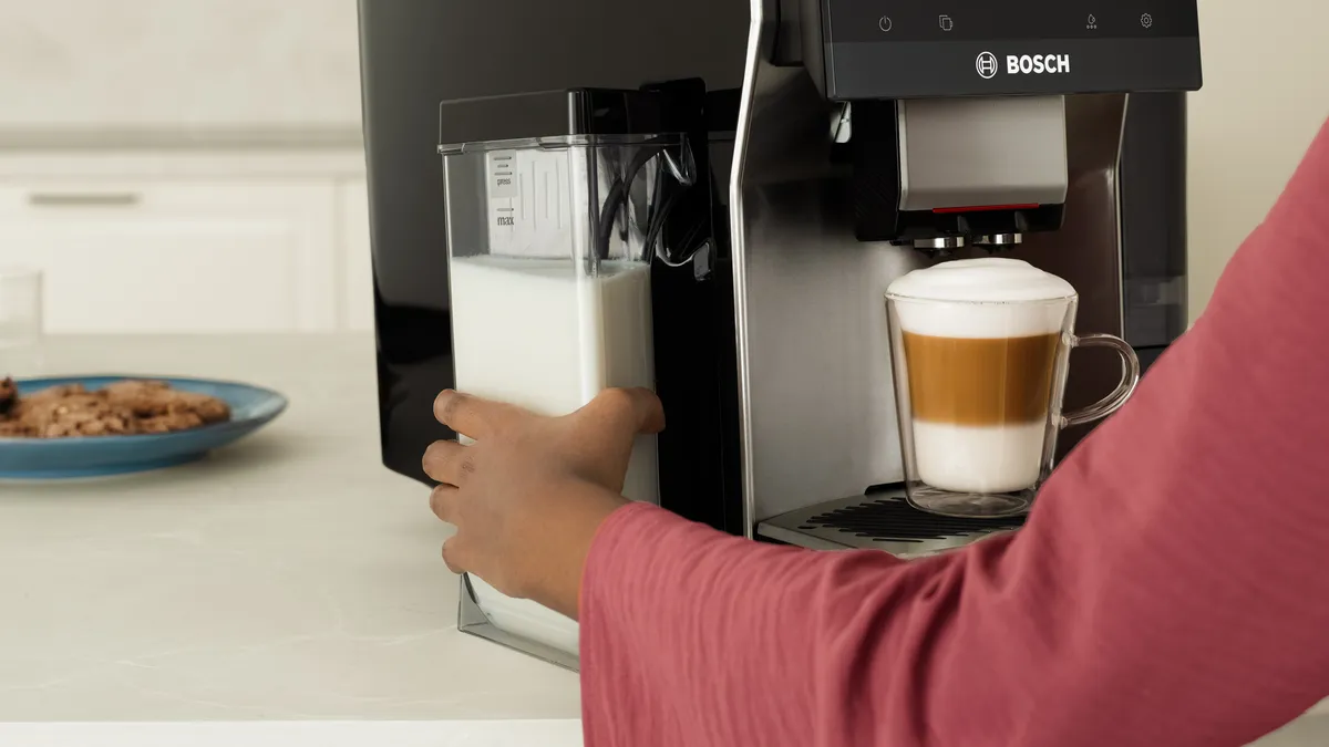 A person in a pink shirt is filling a Bosch coffee machine with milk, while a cup of cappuccino sits on the counter beside a plate of cookies.