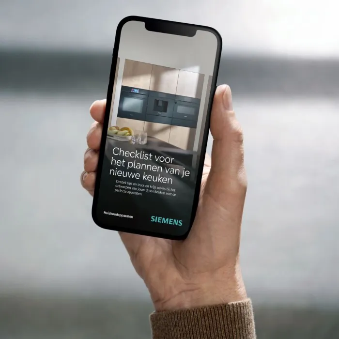 Een persoon houdt een smartphone vast waarop een moderne Siemens keuken met strakke apparaten te zien is, tegen een lichte houten achtergrond.