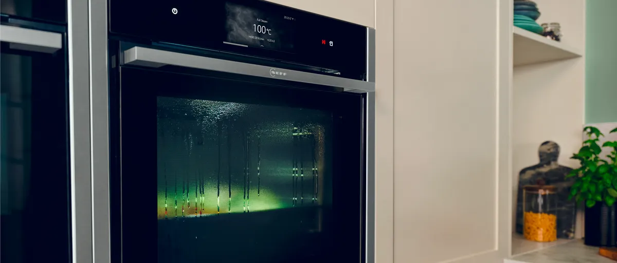 Ein moderner Ofen mit einer Digitalanzeige, die eine Temperatur von 100 °C anzeigt, beschlagenem Glas und im Inneren reflektiertem grünem Umgebungslicht.