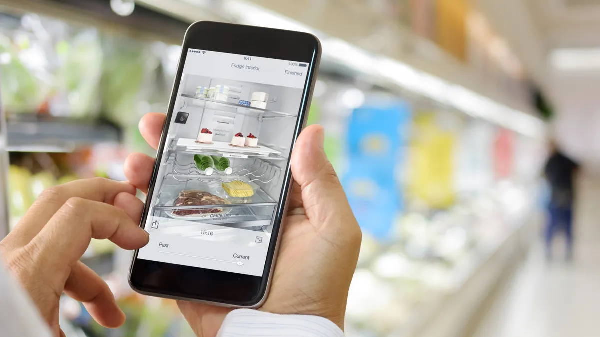 Hände mit einem Smartphone, das einen offenen Kühlschrank mit verschiedenen Speisen im Inneren zeigt.