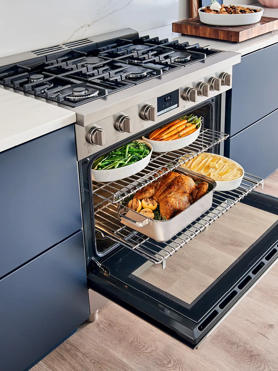 An open oven displays a roasted chicken with vegetables, while a stovetop above features modern gas burners and adjacent bowls of prepared sides.