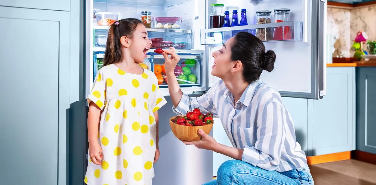 Mutfakta anne ve kız, kızı çilek yiyor, arka planda buzdolabı.