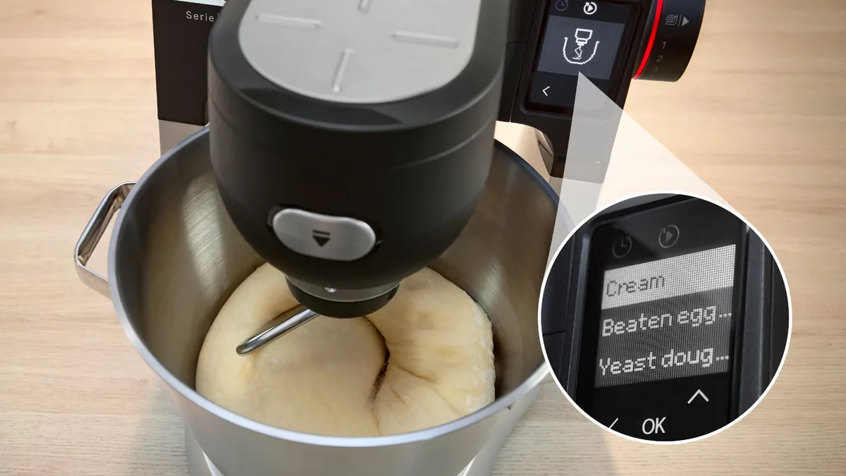 An electric stand mixer is shown in a stainless steel bowl with dough being mixed. The mixer is equipped with a digital display that indicates options such as "Cream," "Beaten egg," and "Yeast dough."