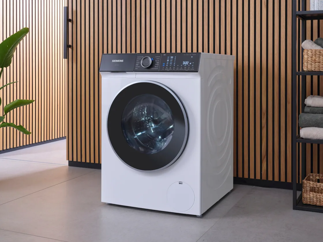 A Siemens iQ500 washing machine, white with a black control panel and door, stands in a room with a slatted wooden wall