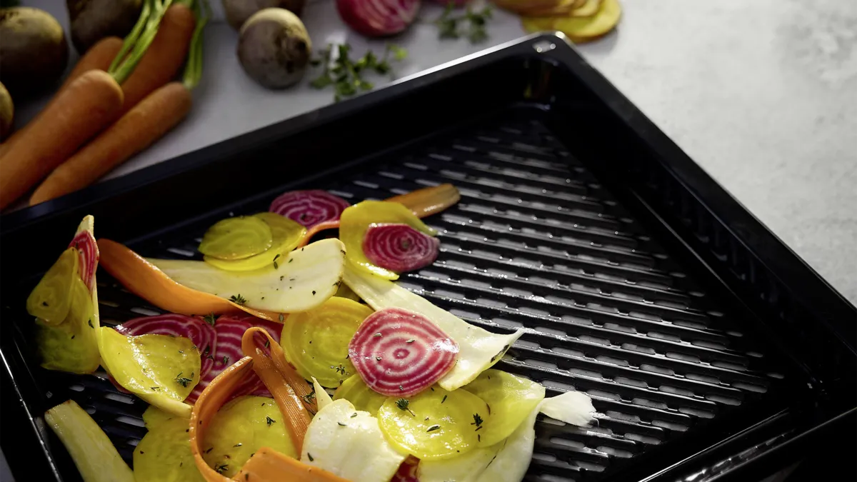 En grillplåt på en köksbänk med skivade färgglada rotfrukter i. I bakgrunden ser man hela rotfrukter.
