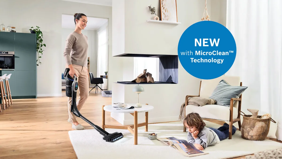 Person vacuums modern living room as child reads by coffee table; promo graphic showcases new tech.