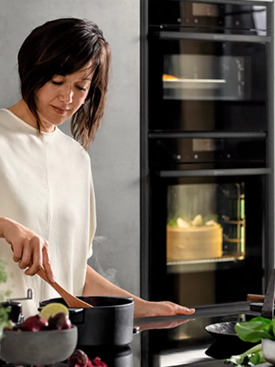 Μια γυναίκα μαγειρεύει σε μια κουζίνα, χρησιμοποιώντας μια κατσαρόλα κι ψήνοντας ένα κέικ στον πίσω φούρνο.
