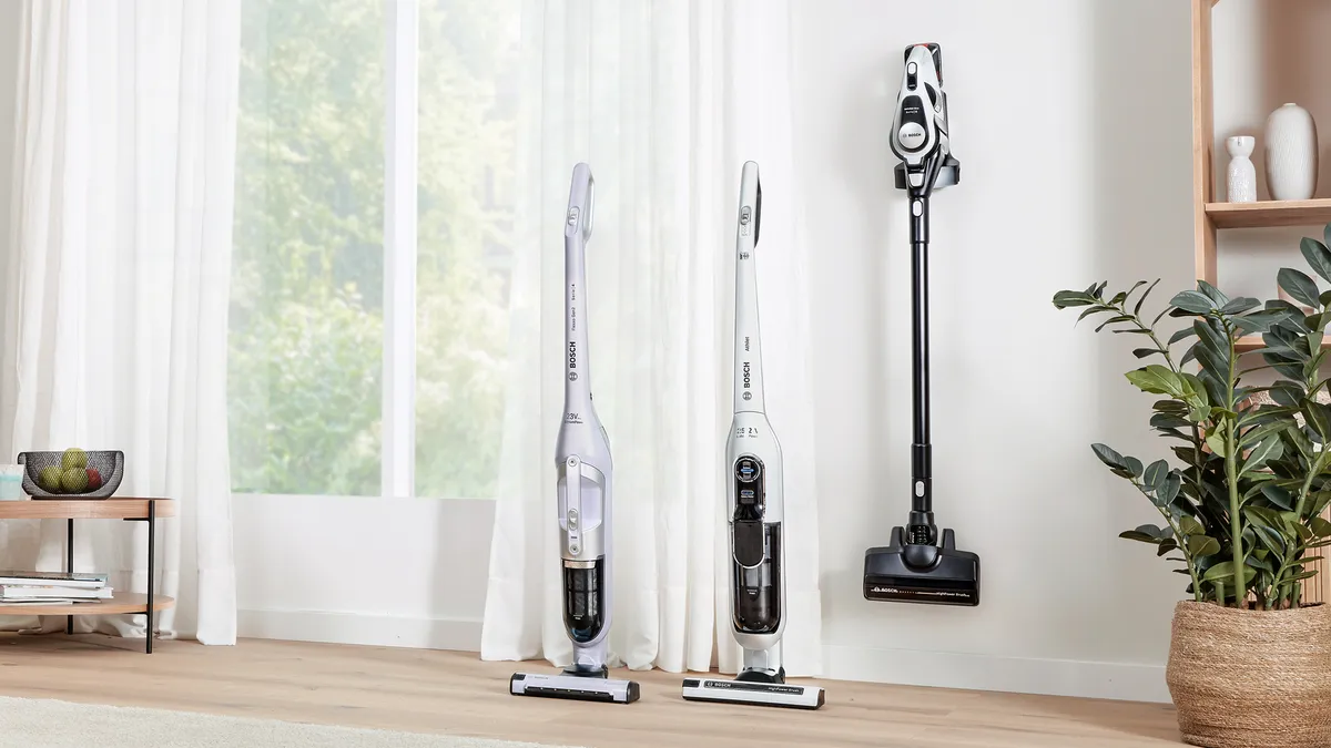 Three cordless vacuums, two standing upright and one mounted on the wall, are positioned alongside the large window of a warm, bright living area.