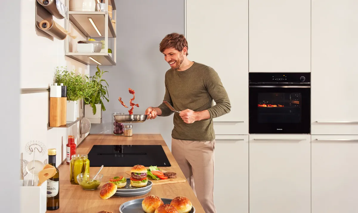 Ein modernes Induktionskochfeld mit einer Pfanne, die Gemüsepatties enthält, einem dampfenden Topf und frisch gebackenem Brot auf einem Holzbrett.