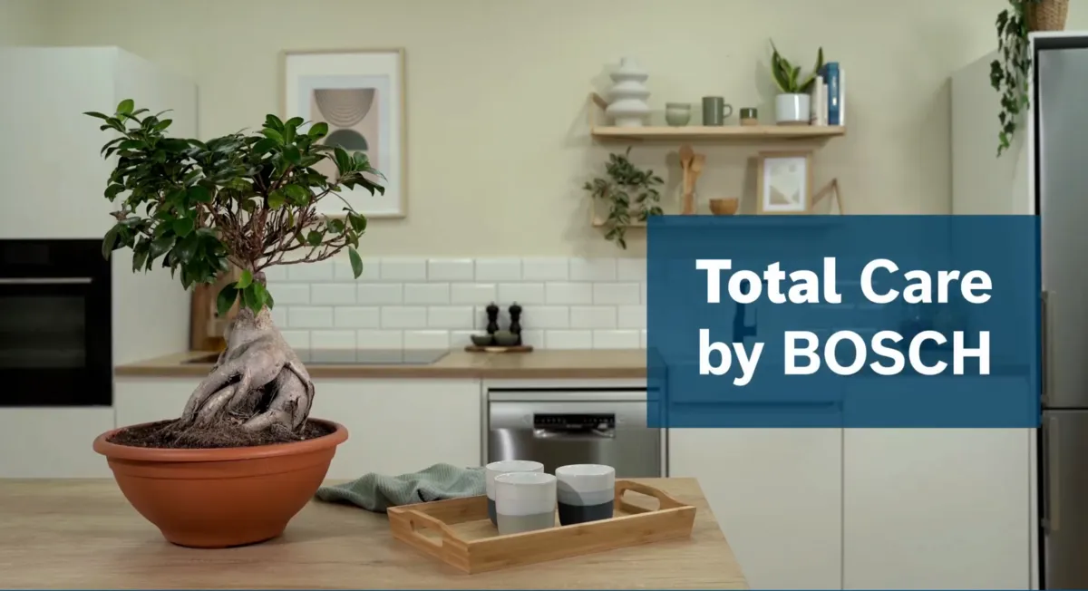 Een keuken met een bonsaiboompje op een kookeiland en een spandoek ervoor waarop reclame wordt gemaakt voor de Bosch Total Care-diensten.