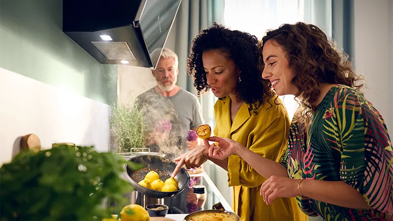 Two women in a bright kitchen are cooking together, with one holding a grilled lemon and steam rising from a pan of potatoes.