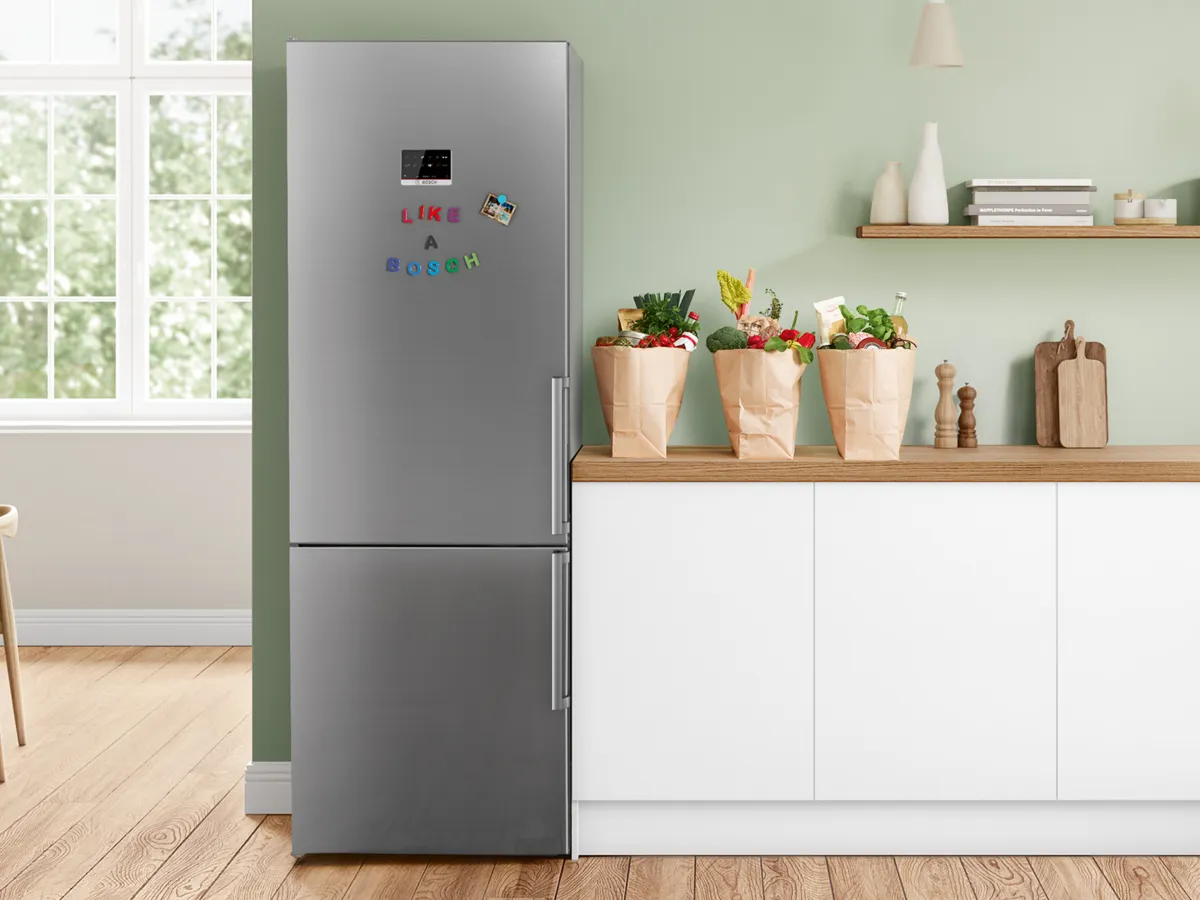 Réfrigérateur Bosch inox avec aimants colorés, à côté de sacs de courses posés sur un plan de travail.