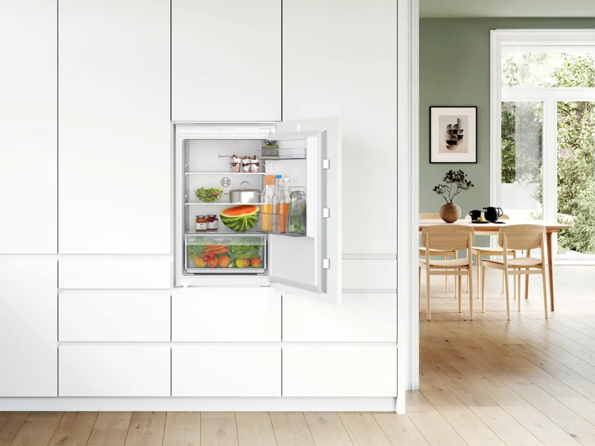 Petit frigo encastré ouvert, rempli de fruits, légumes et boissons, dans une cuisine moderne.