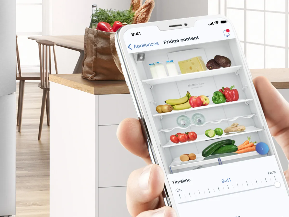 Une main tient un smartphone affichant un inventaire numérique du réfrigérateur, tandis qu'une cuisine avec des produits d'épicerie et une table en bois se trouve en arrière-plan.