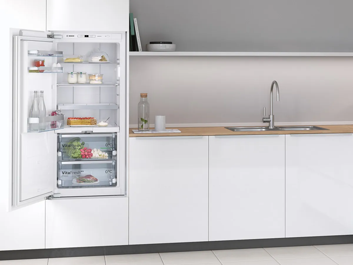 Réfrigérateur encastrable ouvert dans une cuisine blanche moderne avec fruits et produits frais.