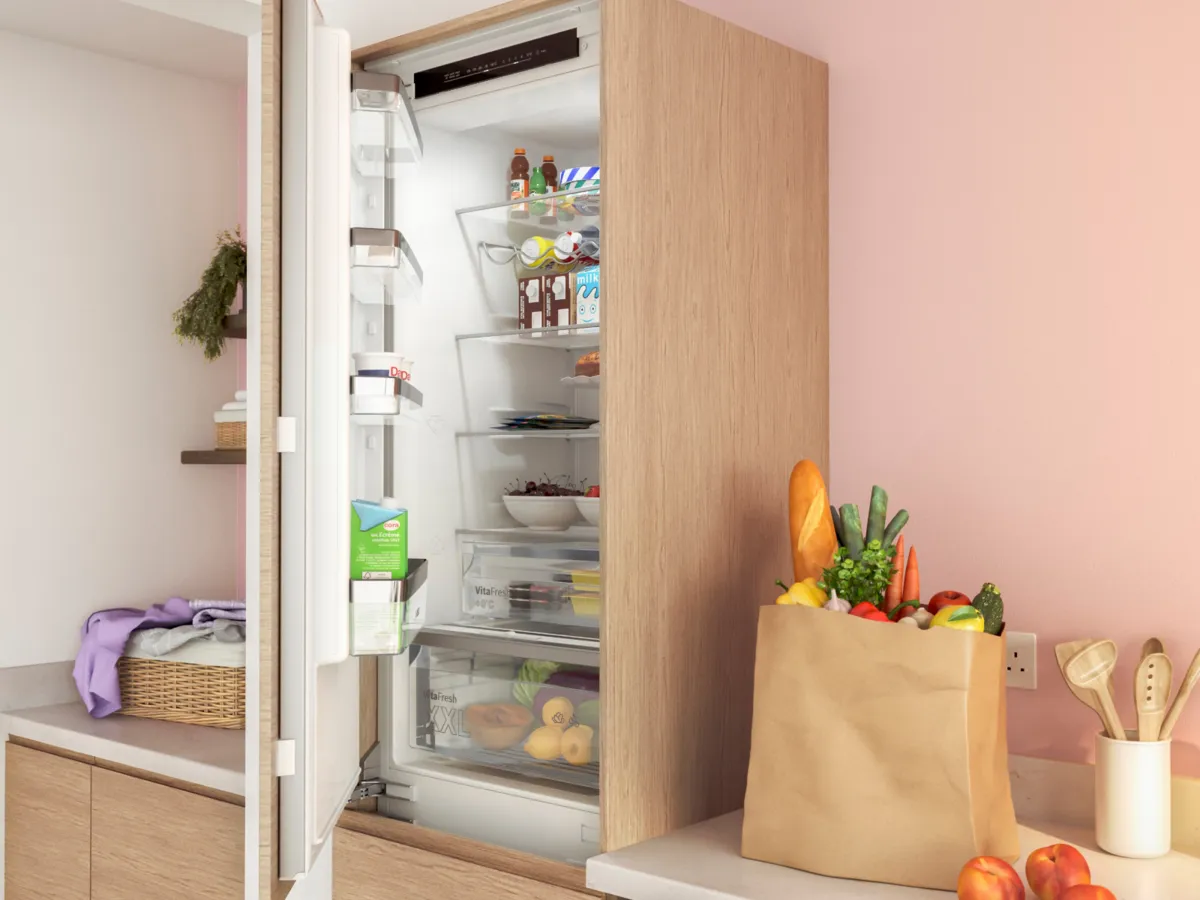 Réfrigérateur encastré ouvert dans une cuisine bois et rose, avec un sac de courses rempli à côté.