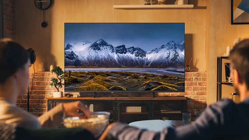 Cozy live room with large TV displaying kary dağ manzara, tuğla duvar ve ahşap raflar.