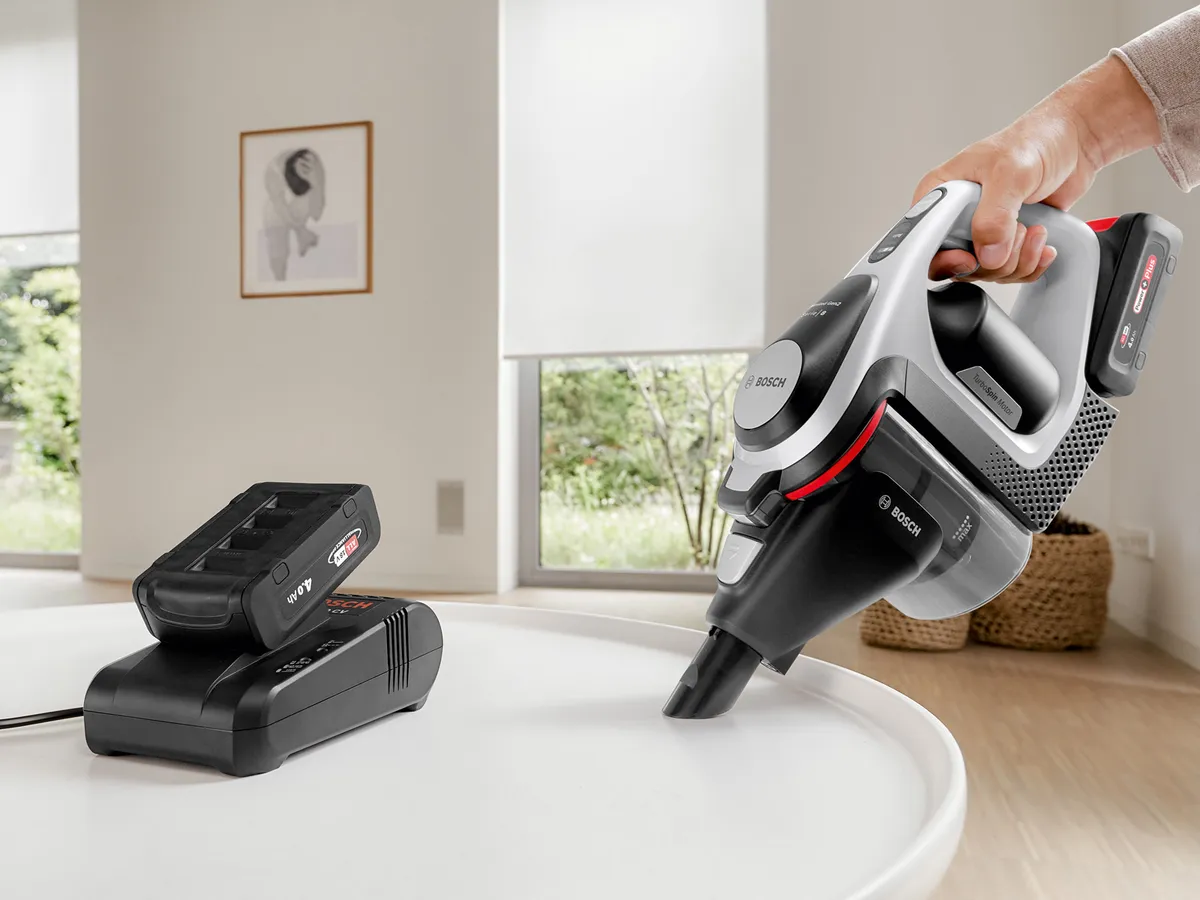 Eine Person reinigt eine Oberfläche mit dem Bosch Unlimited Staubsauger. Daneben ist ein Akku in einer Ladestation aufgestellt.