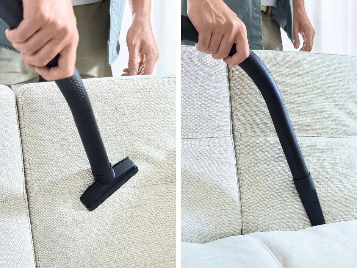 Nahaufnahme einer Hand, die mit der Fugendüse Schmutz von schwer erreichbaren Stellen auf der Couch entfernt. Die Polsterdüse wird verwendet, um die Polster der Couch abzusaugen.