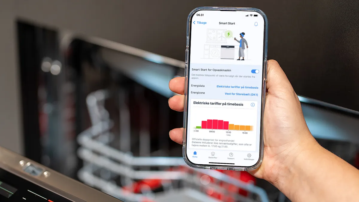 En person holder en smartphone med "Smart Start"-funktionen for en opvaskemaskine, med en farverig energi-graf.
