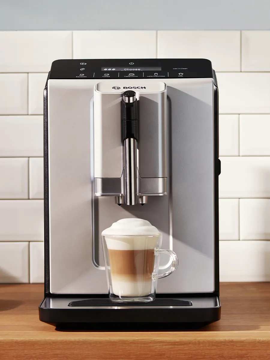 آلة قهوة بوش أوتوماتيكية بالكامل ذات لون فضي وأسود تصنع مشروب قهوة متعدد الطبقات في كوب زجاجي شفاف، وتظهر خلفها بلاط أبيض. 