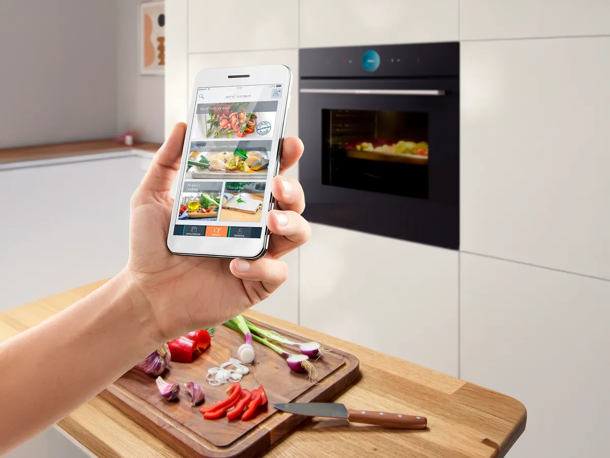 Mão segurando um smartphone com receitas de comida, fundo de cozinha com vegetais numa tábua de cortar.