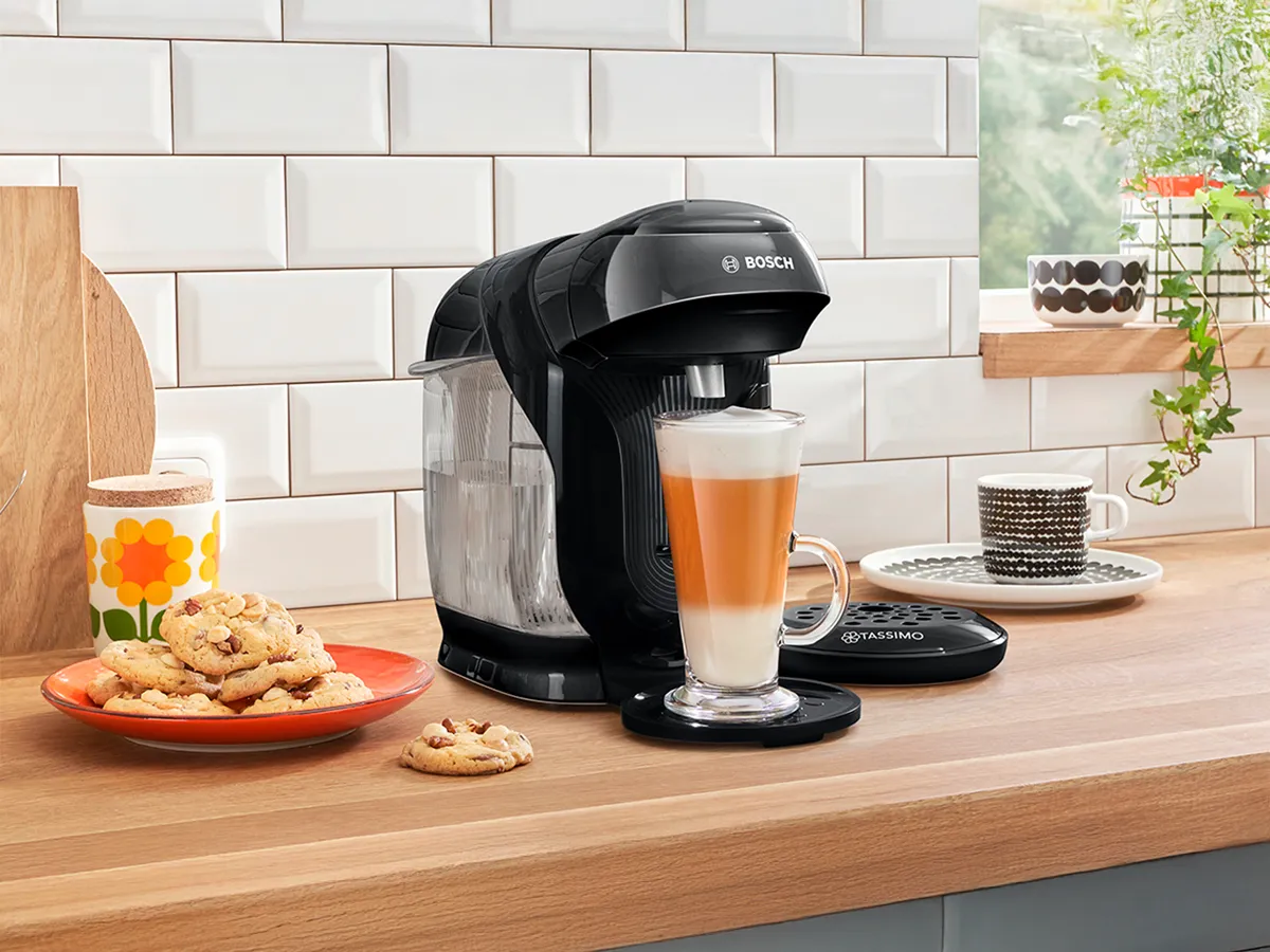 TASSIMO STYLE friendly brewing a Latte Macchiato on a kitchen worktop and a plate of cookies next to it.