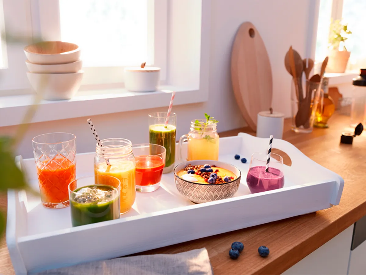 Bandeja com smoothies coloridos e uma tigela de frutas em uma cozinha iluminada, apresentando opções de bebidas frescas e saudáveis.