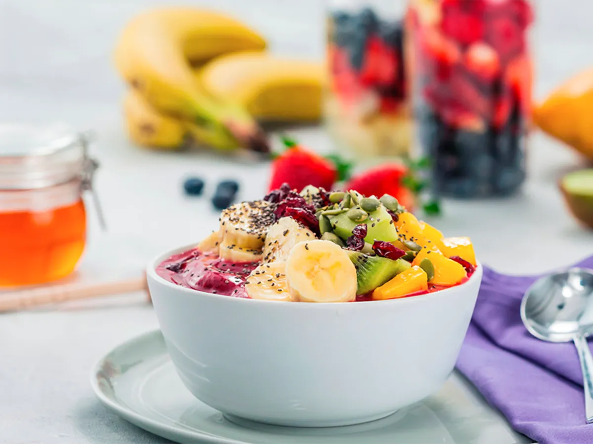 Tigela de smoothie colorida coberta com frutas frescas, sementes e mel, rodeada por bananas e frutas vermelhas para um café da manhã vibrante.