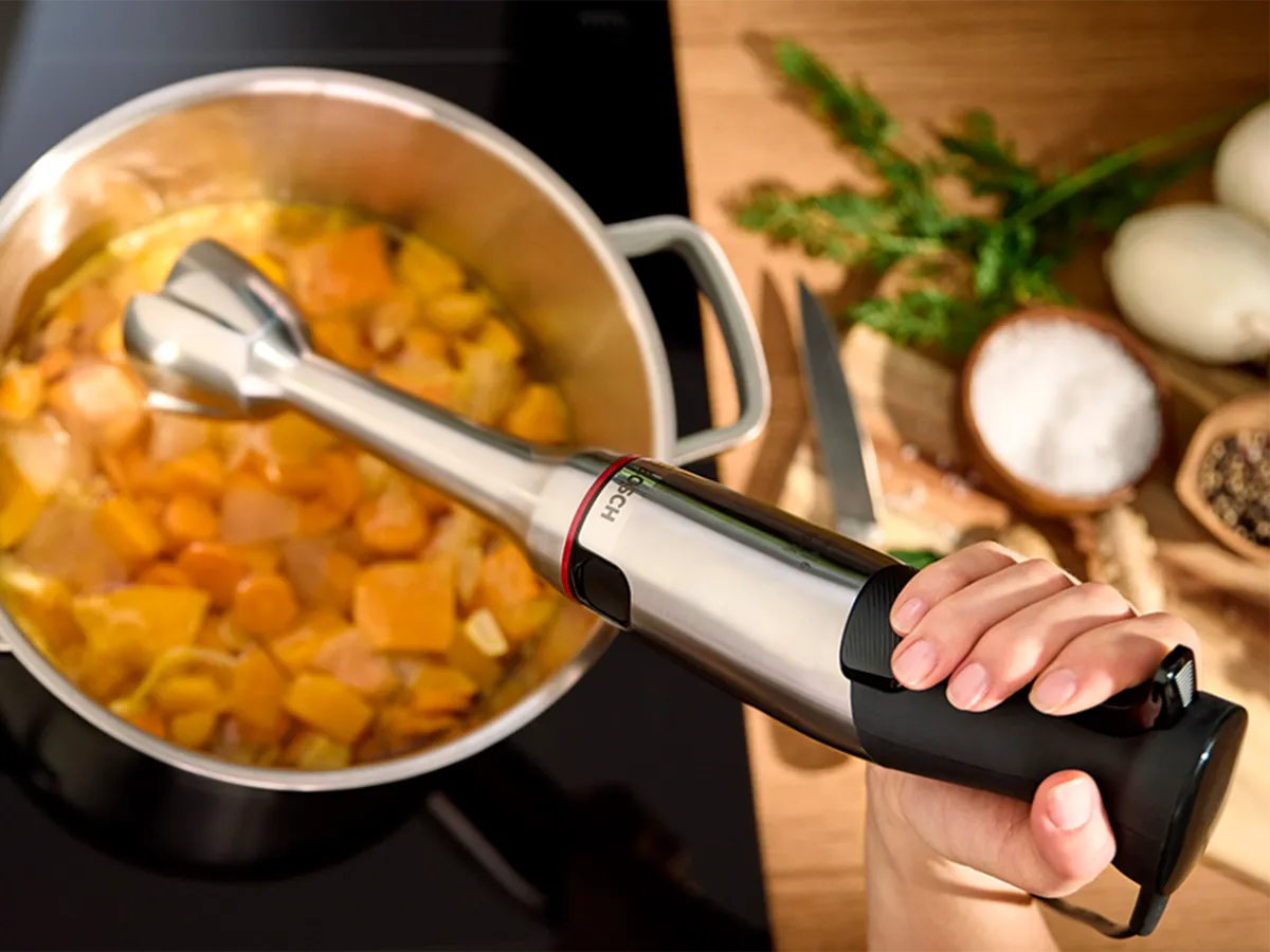Uma mão segura um liquidificador de imersão sobre uma panela de legumes recém-cozidos, pronta para fazer uma sopa cremosa.