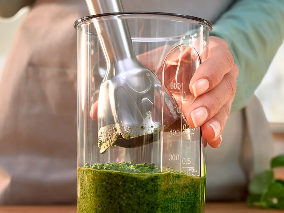 Um liquidificador de imersão paira sobre um copo cheio de pesto verde recém-preparado, que está em uma bancada de madeira.
