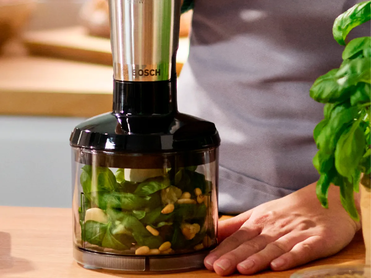 Uma pessoa segura um liquidificador de mão Bosch, usando o acessório picador para cortar vegetais.