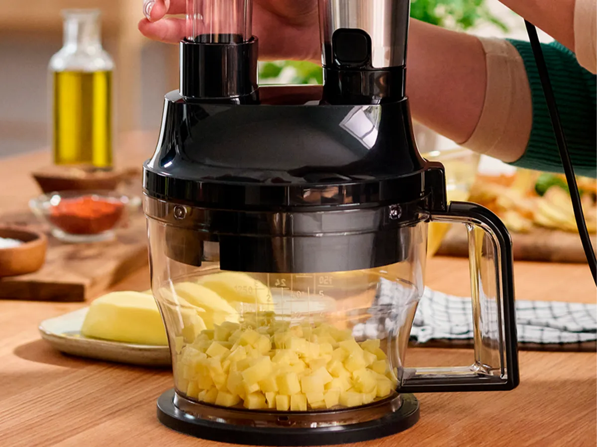 Uma mão opera um liquidificador equipado com o acessório cortador em cubos, que repousa sobre uma bancada de madeira e está cortando batatas em cubos.