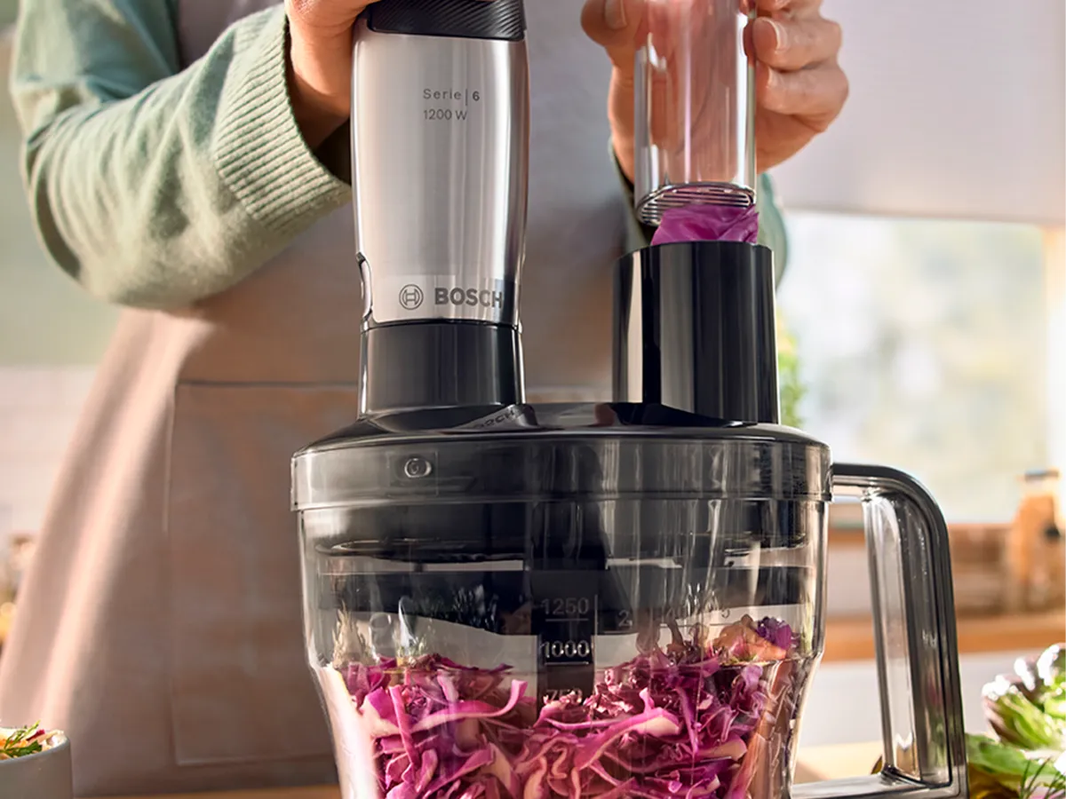 Uma pessoa com avental usa um processador de alimentos para ralar repolho-roxo.