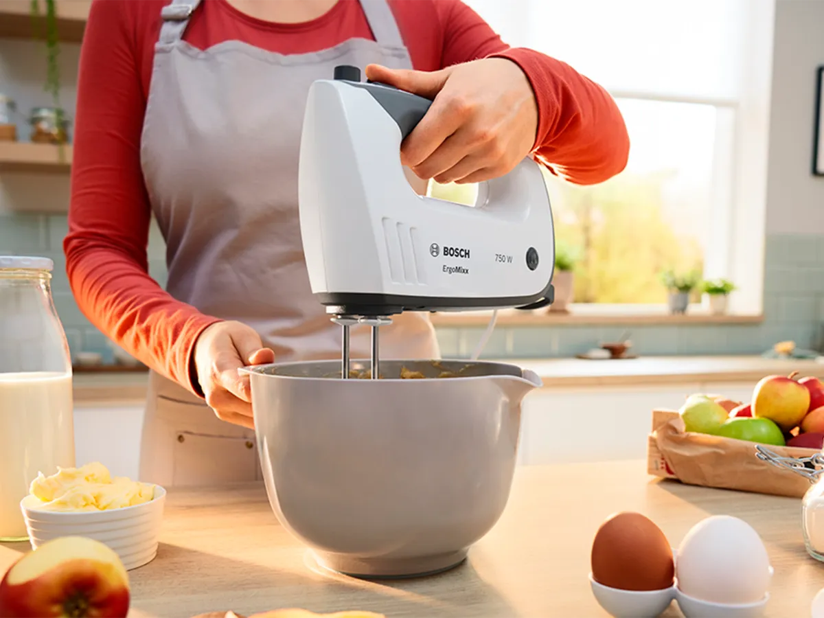 Uma pessoa usa um liquidificador de mão Bosch para bater o conteúdo de uma tigela branca.