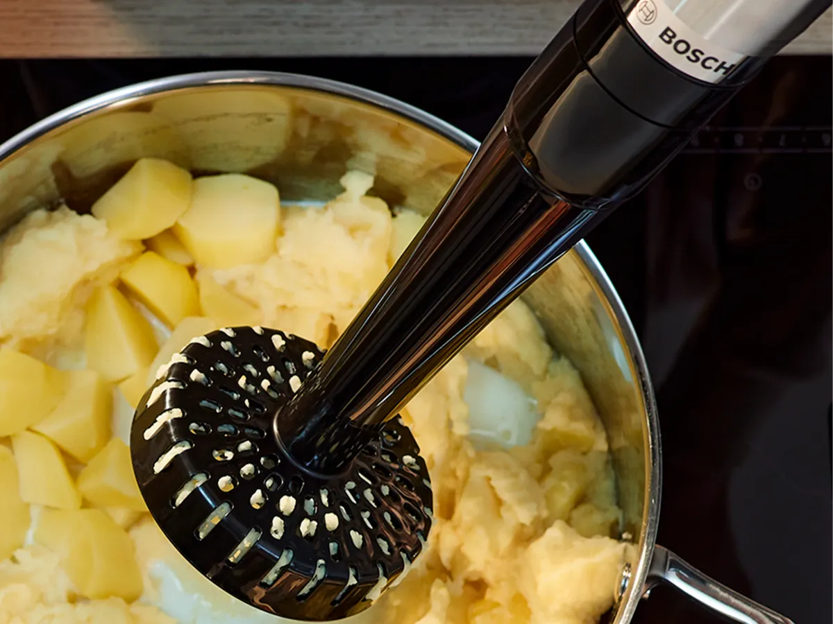 Um liquidificador com o acessório de amassar paira acima de uma panela com cubos de batata recém-cozidos.