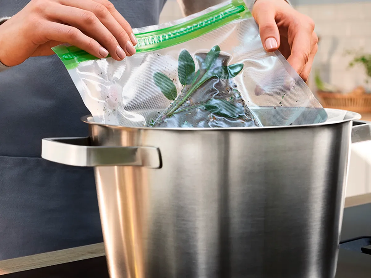 As mãos seguram um saco de verduras selado a vácuo sobre uma panela, preparando-se para cozinhá-las sous vide.
