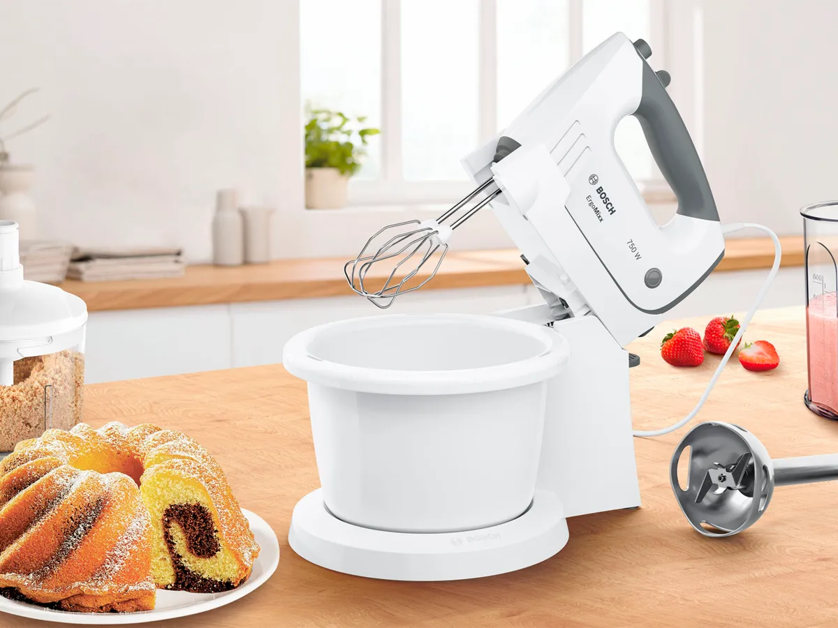 Uma batedeira Bosch com batedeiras e um processador de alimentos, ao lado de um bolo e morangos, numa bancada de madeira de uma cozinha luminosa.