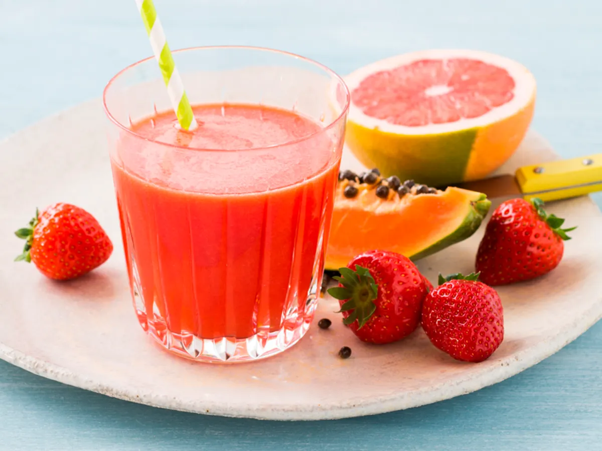 Um copo de suco vermelho fresco com um canudo, cercado por morangos, fatias de mamão e toranja em um prato de cor clara.