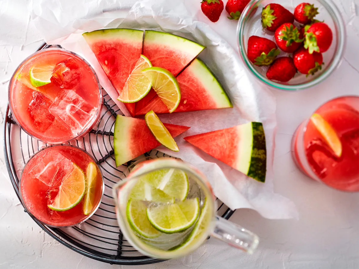 Um vibrante flat lay de refrescantes cocktails de melancia com lima, acompanhados por melancia fatiada e morangos frescos.