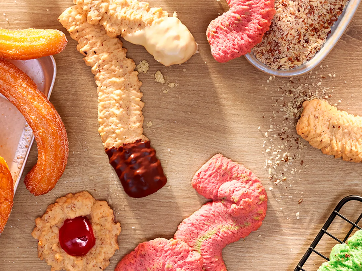 Biscoitos coloridos variados e churros numa superfície de madeira, alguns mergulhados em chocolate e cobertos com nozes.