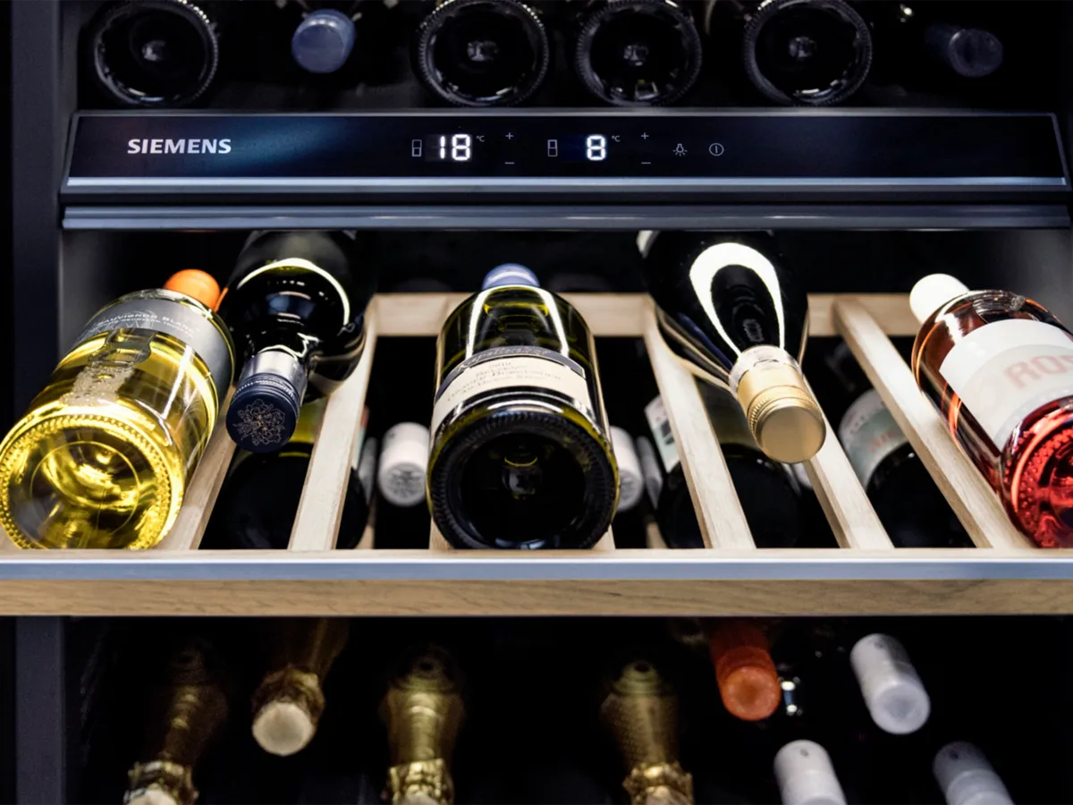 Close-up of a wine cooler with wooden racks holding various bottles, displaying a Siemens temperature control panel showing two temperatures.