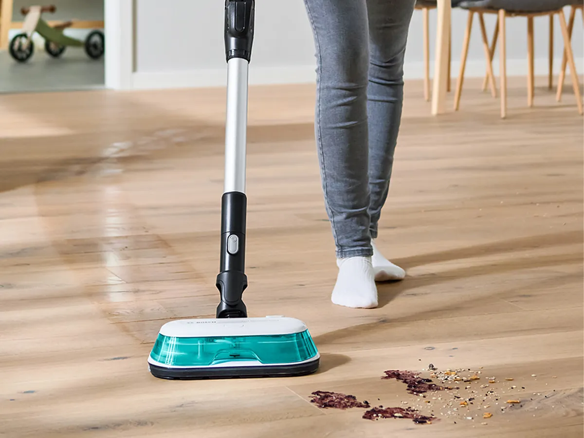 Oseba v sivih hlačah in bosih nogah uporablja električni čistilec tal na lesenem podu s raztresenimi drobtinami.