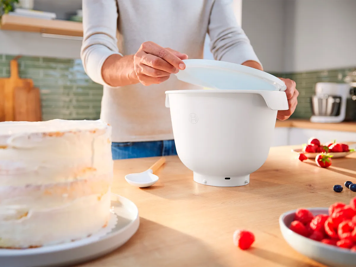 Mulher a colocar a tampa numa taça de plástico Flex Bowl ao lado de um pão de ló e de um patê de fruta colocados numa banca de cozinha.