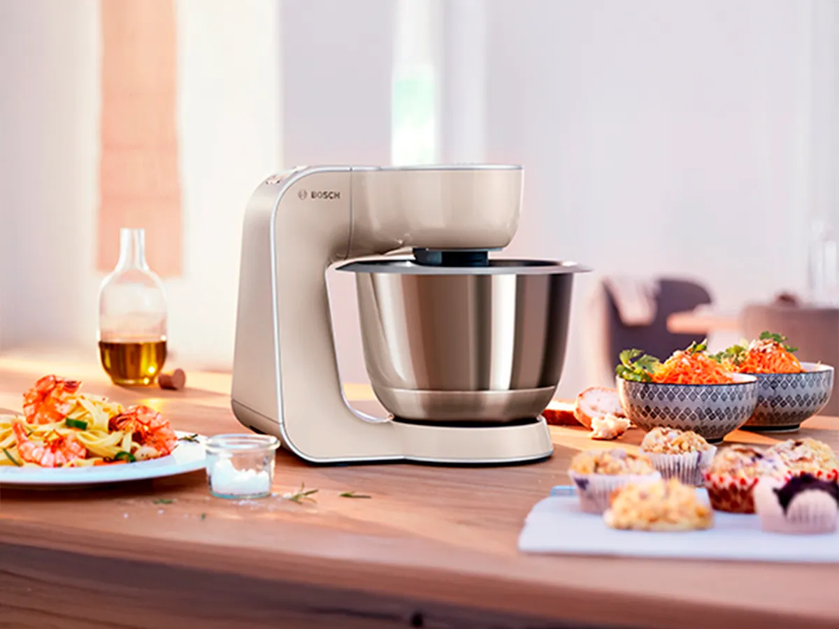 Batedeira Bosch moderna com taça brilhante, rodeada de pratos variados sobre mesa de madeira, criando um ambiente vibrante.