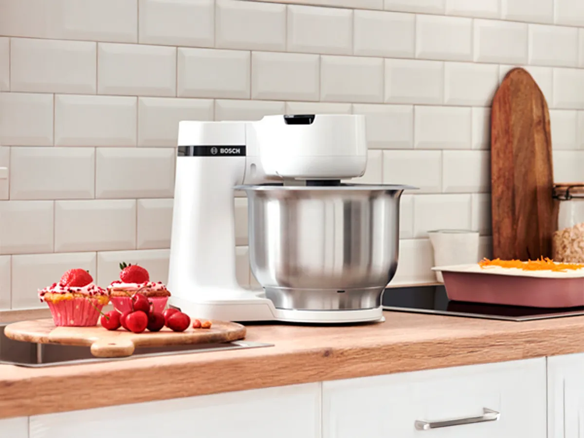 Batedeira Bosch branca com taça inox sobre balcão de madeira, rodeada de queques e ingredientes para pastelaria.