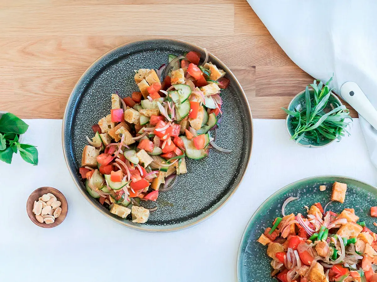 Vista de cima de uma salada com pepinos, tomates, croutons e verduras, servida num prato com padrão salpicado.