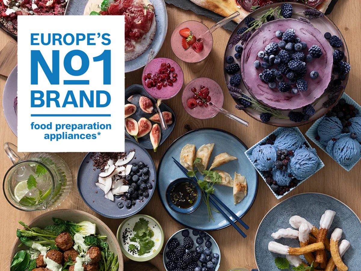 Aparelhos de preparação de alimentos, utensílios de cozinha, produtos frescos e um livro intitulado "Europe's No1 Brand" em uma mesa de madeira. 