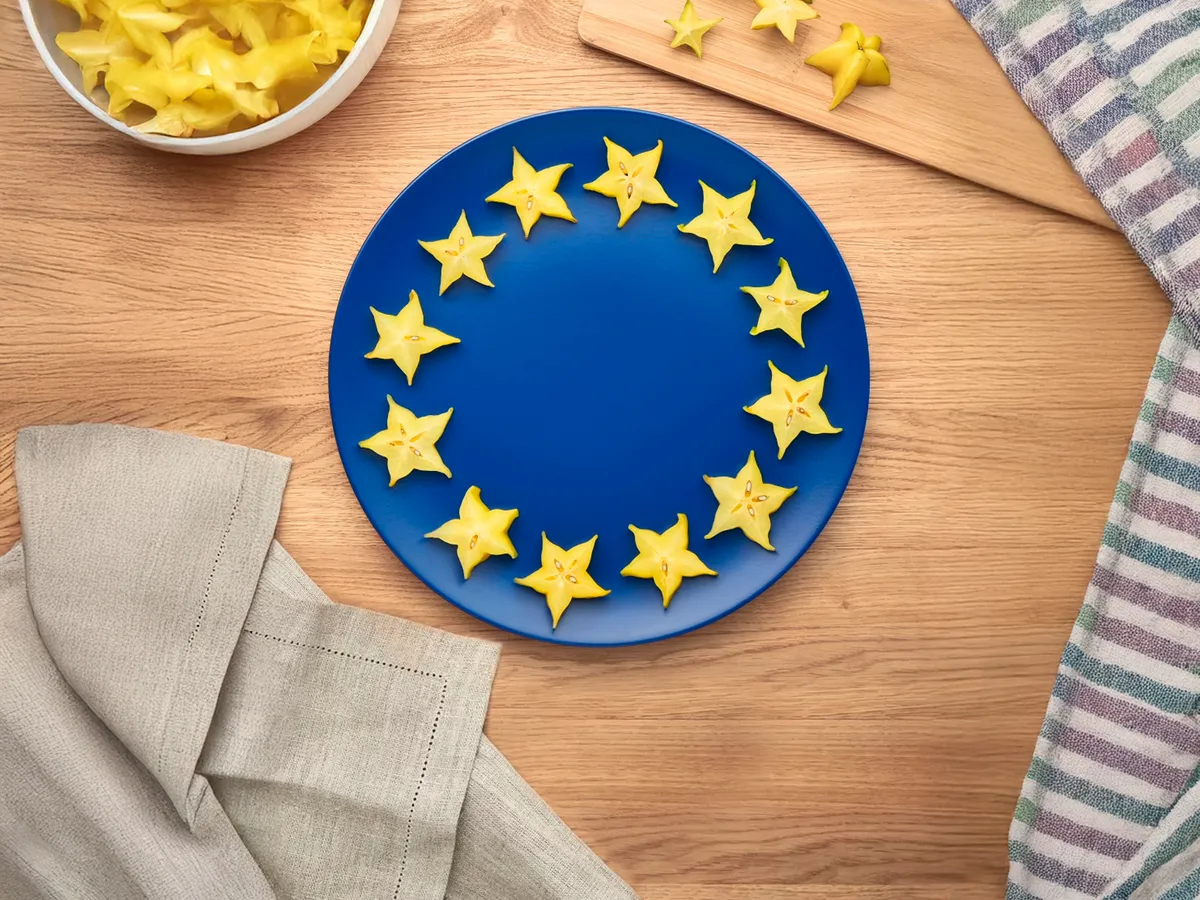Um prato azul decorado com frutos amarelos em forma de estrela, dispostos como a bandeira da União Europeia, colocado numa mesa de madeira com um pano e uma toalha às riscas.